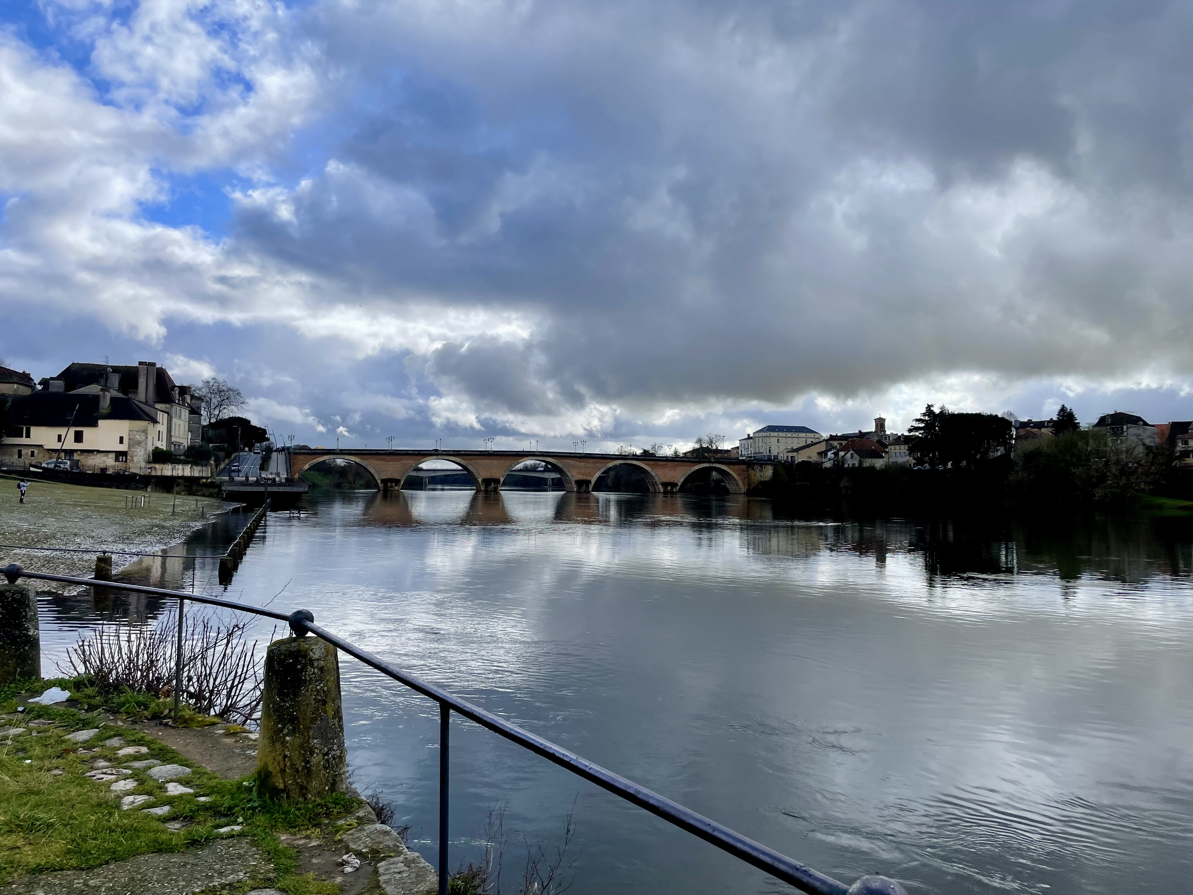 View from the edge of the promenade along the Dordogne River toward the old stone bridge in the distance. This bridge connects the two halves of Bergerac, and is used by cars, buses, bicycles, small electric scooters, and pedestrians alike. In the photo, the sky above has puffy clouds with bits of blue peeking through, and below the river is like a mirror, reflecting city, bridge, and sky perfectly.
